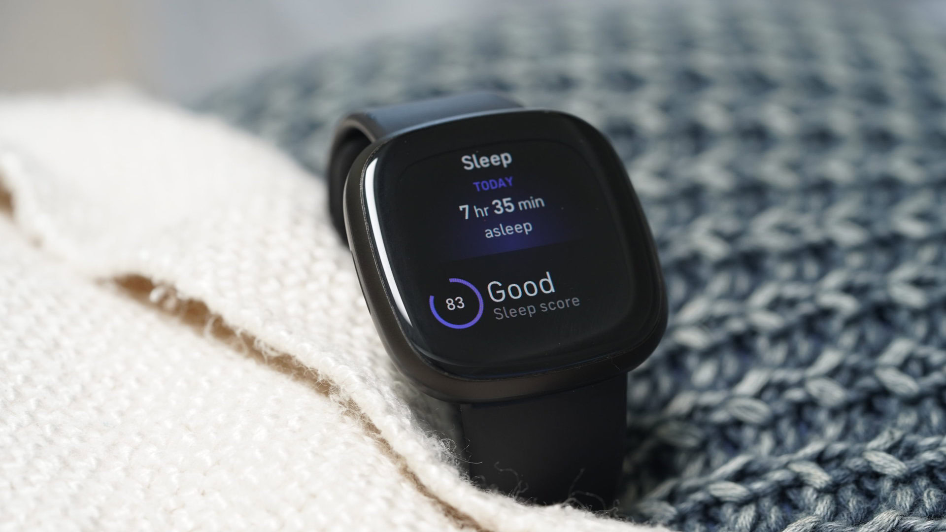 Um Fitbit Versa 3 repousa em uma pilha de roupas de cama, exibindo os dados de sono de um usuário.