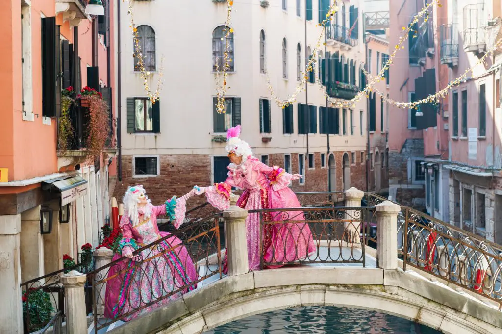 Duas pessoas em elaborados trajes rosa de estilo do século XVIII e perucas brancas posando em uma pequena ponte em arco sobre um canal em Veneza, cercado por edifícios históricos e luzes festivas de cordas.