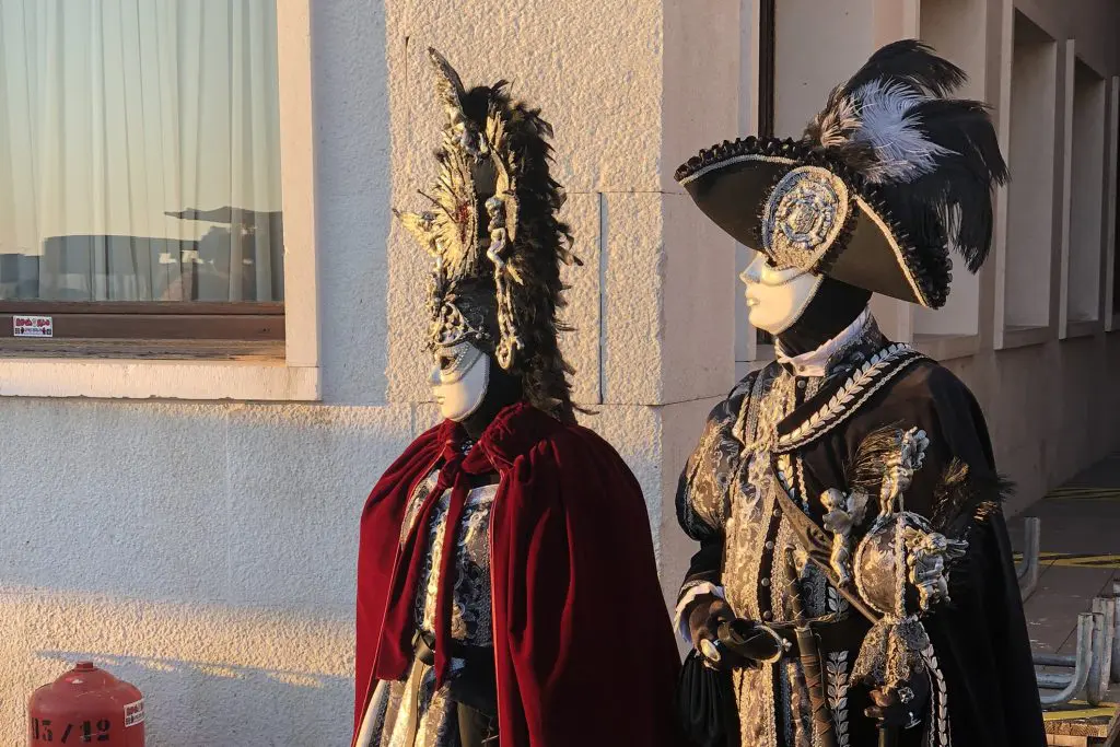 Duas pessoas em figurinos elaborados de carnaval veneziano com máscaras ornamentadas, penas e bordados em estilo barroco, de pé contra uma parede de construção de cor clara durante o pôr do sol
