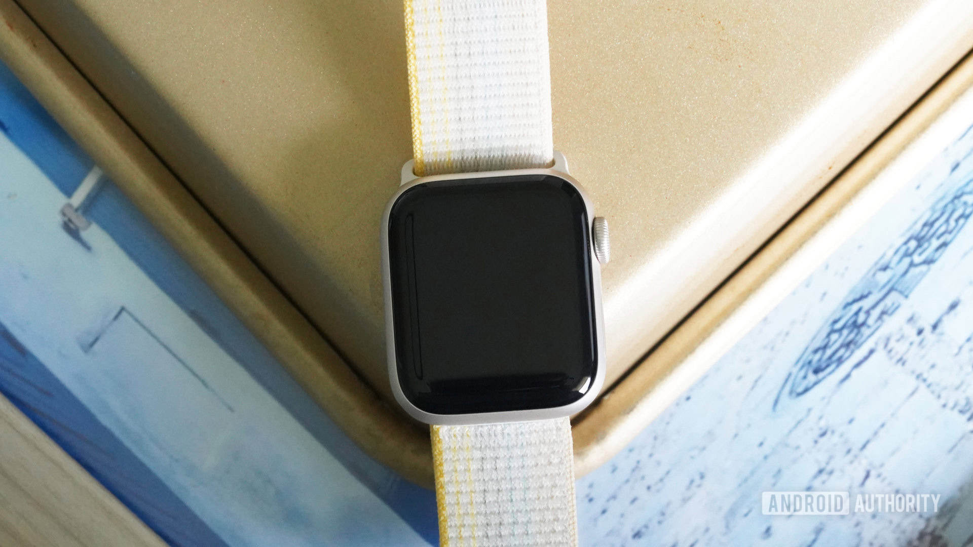 Tela em branco do Apple Watch SE 2 Um Apple Watch SE 2 repousa sobre uma superfície dourada e azul com a tela desligada.