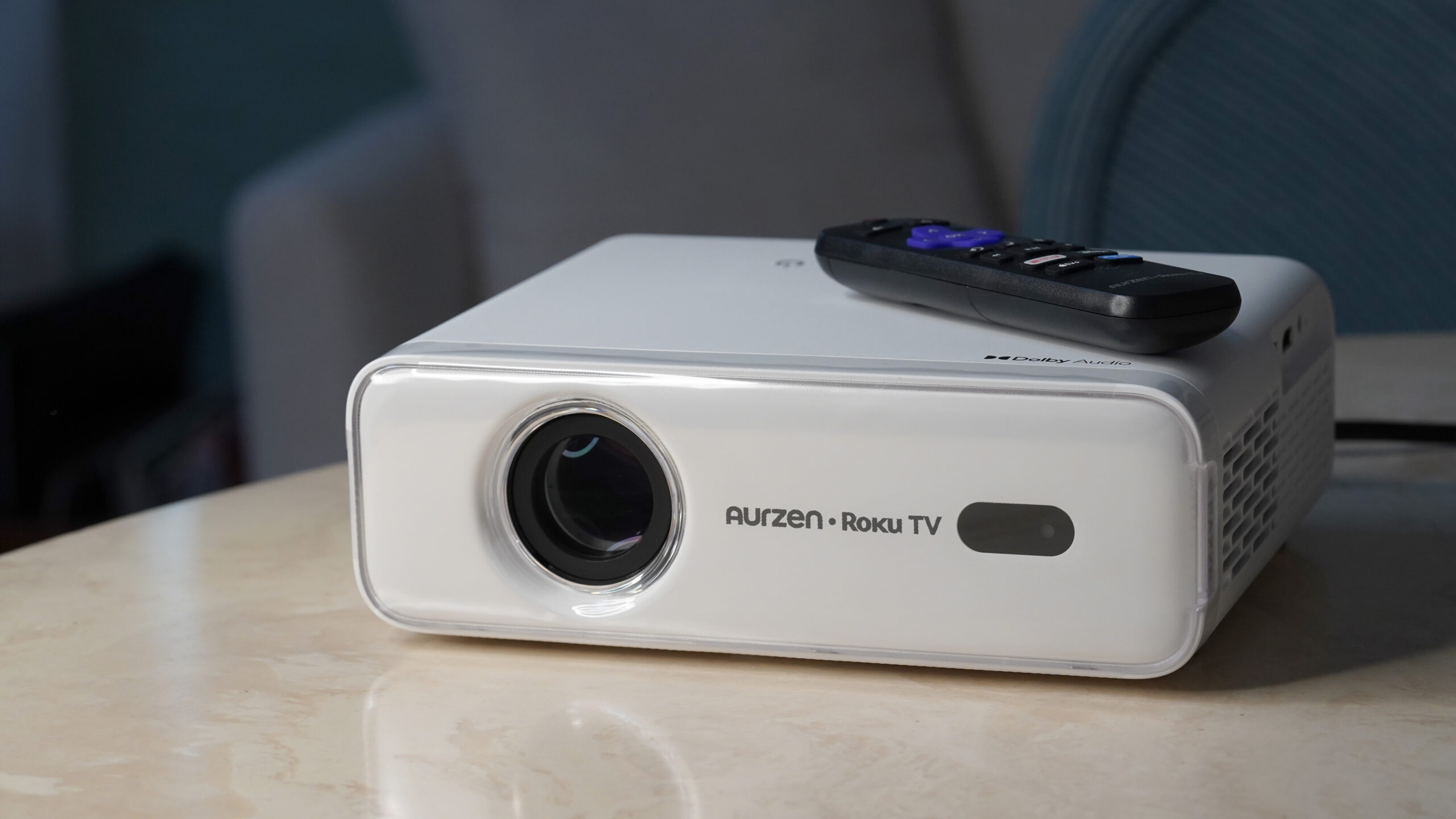 Projetor Roku TV TV EAZZE D1R Um projetor Aurzen Roku TV EAZZE D1R repousa sobre uma superfície de mármore com seu controle remoto incluído.