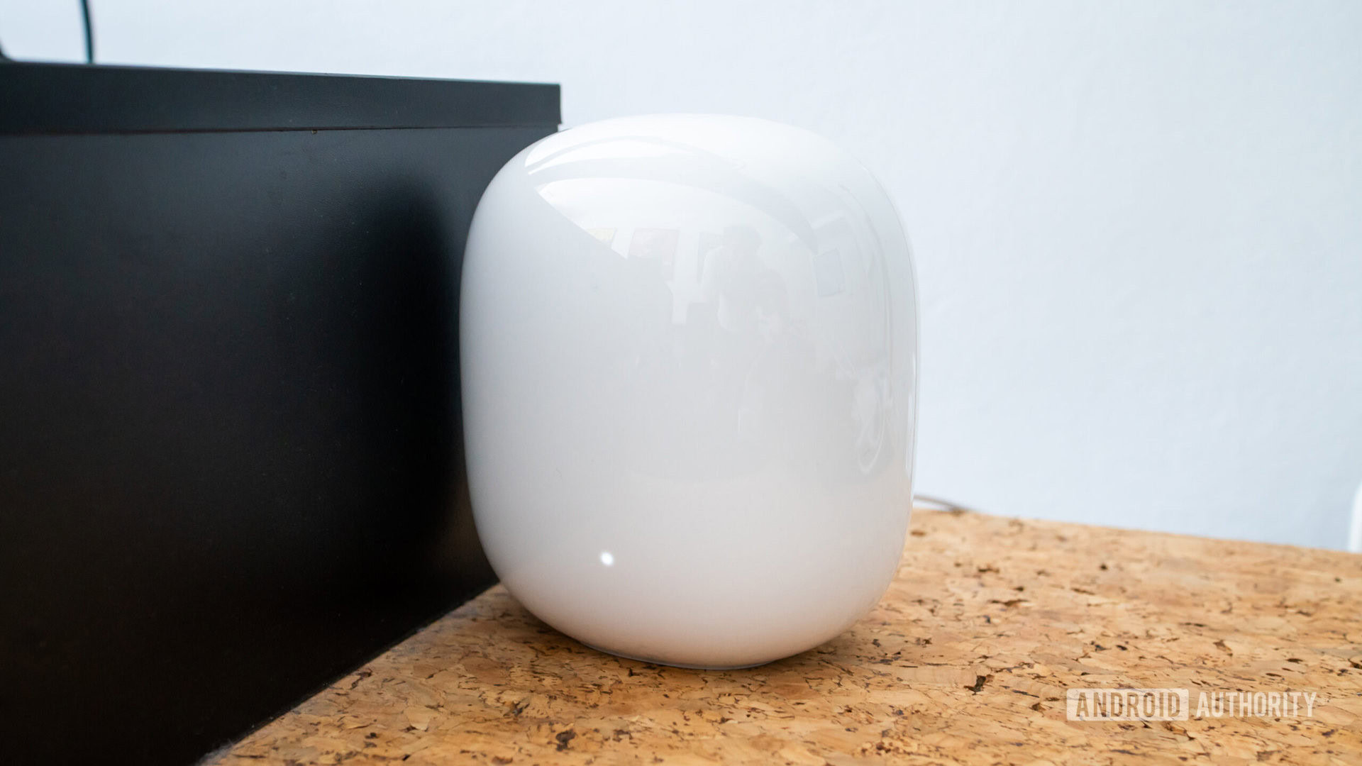 Nó Google Nest Wi-Fi Pro em uma mesa de cortiça ao lado de um suporte de monitor de madeira preto Nó Google Nest Wi-Fi Pro em uma mesa de cortiça ao lado de um suporte de monitor de madeira preta