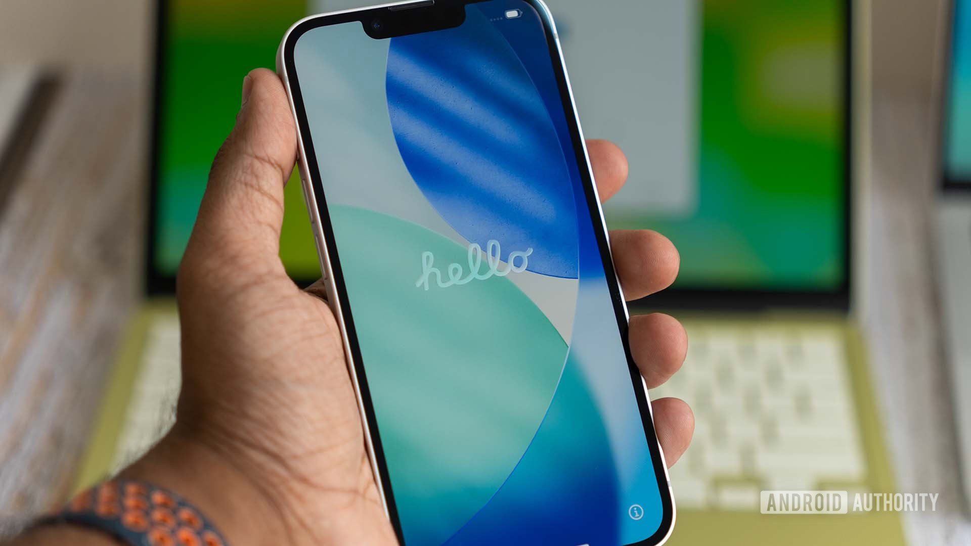 Apple iPhone 17e mostrando a tela de inicialização de Olá durante a inicialização, com o MacBook Neo em segundo plano